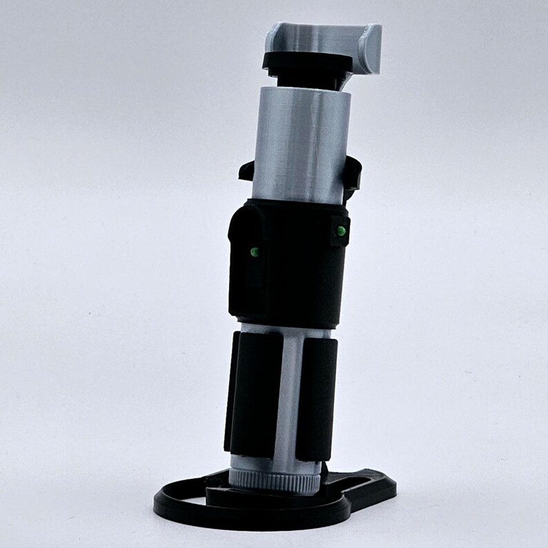 May include: A silver and black lightsaber prop with a green light on the side, mounted on a black stand.