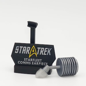 Może przedstawiać: Czarny stojak ekspozycyjny z napisem STAR TREK STARFLEET COMMS EARPIECE oraz żółtym symbolem delta. Obok znajduje się szara słuchawka douszna z cylindrycznym, żebrowanym elementem połączonym z małą, półprzezroczystą, wygiętą końcówką douszną.