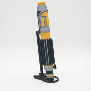 May include: A cylindrical prop handle featuring a gray and black body with bright yellow accents. The handle has a ribbed grip section, a top emitter with yellow detailing, and is mounted vertically on a black plastic display stand.