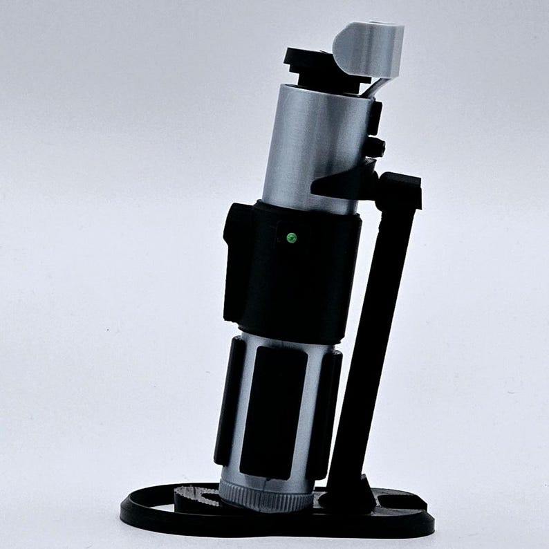 May include: A black and silver lightsaber prop with a green light on the side, mounted on a black stand.