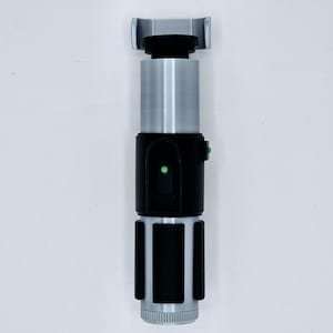 May include: A black and silver lightsaber prop with a green light. The lightsaber has a black handle with a silver blade and a black grip.