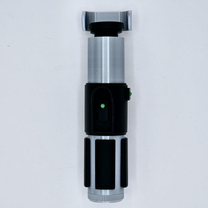 May include: A black and silver lightsaber prop with a green light. The lightsaber has a black handle with a silver blade and a black grip.