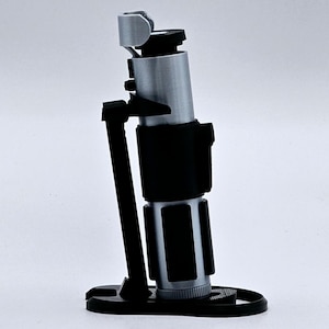 May include: A silver and black lightsaber prop with a stand. The lightsaber is made of plastic and has a detailed design. The stand is black and has a simple design.
