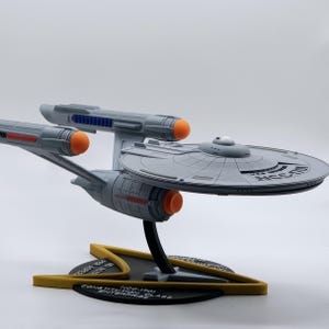 Puede incluir: Un modelo 3D impreso en gris del USS Enterprise NCC-1701, la nave espacial de Star Trek. El modelo está sobre una base negra con un emblema de la Flota Estelar en oro y el texto "NCC-1701 CONSTITUTION CLASS ENTERPRISE".