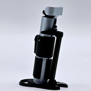 May include: A silver and black lightsaber replica with a green light on the side, displayed on a black stand.