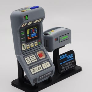 May include: A gray Star Trek TR-560 Tricorder VI model with a medical module. The Tricorder has a flip-up screen, buttons, and a red button. The medical module has a green display. The base has the Star Trek logo.