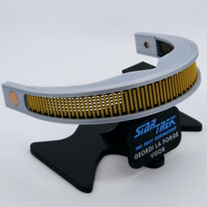 Può includere: Replica del visore di Geordi La Forge di Star Trek: The Next Generation, grigio e giallo, su un supporto nero. Il visore curvo presenta barre gialle verticali. La base riporta la scritta "Geordi La Forge Visor".