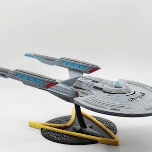 May include: A grey model of the U.S.S. Enterprise NCC-1701-G starship from Star Trek, with blue and red accents. The model is mounted on a black and yellow stand.
