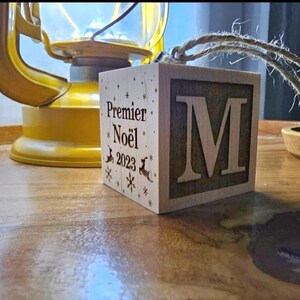 May include: A wooden block ornament with the text "Premier Noël 2023" and the letter "M" on the front. The ornament has a twine hanger.