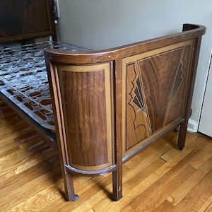 Puede incluir: Un marco de cama de madera antigua con cabecero y piecero. El cabecero presenta un diseño geométrico decorativo con tonos de madera marrón oscuro y claro. El marco de la cama tiene una base de muelles metálicos y está sobre ruedas pequeñas.