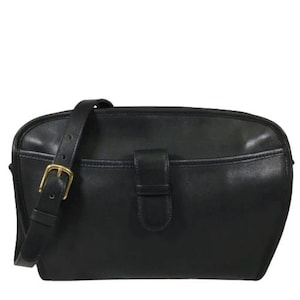 May include: A black leather crossbody bag featuring a smooth finish, a front slip pocket with a central leather tab closure, and an adjustable shoulder strap secured by a brass-toned metal buckle. The bag has a rounded top edge and a structured shape.