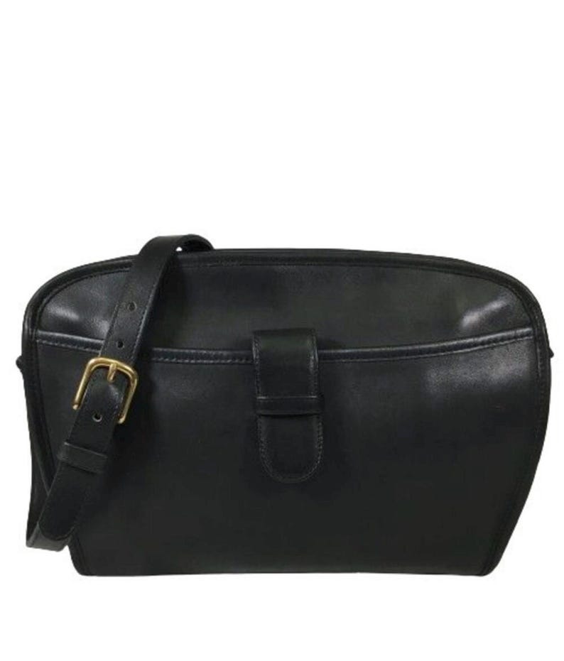 May include: A black leather crossbody bag featuring a smooth finish, a front slip pocket with a central leather tab closure, and an adjustable shoulder strap secured by a brass-toned metal buckle. The bag has a rounded top edge and a structured shape.