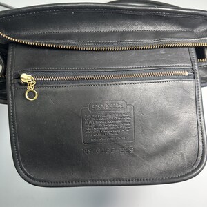 May include: A black leather bag featuring a zippered front pocket with a gold-toned circular pull. The front flap is embossed with the Coach logo and a text block detailing the natural characteristics of the leather, along with the serial number 0486-229.