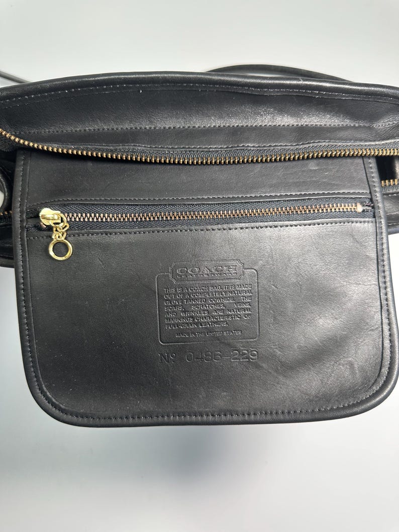 May include: A black leather bag featuring a zippered front pocket with a gold-toned circular pull. The front flap is embossed with the Coach logo and a text block detailing the natural characteristics of the leather, along with the serial number 0486-229.