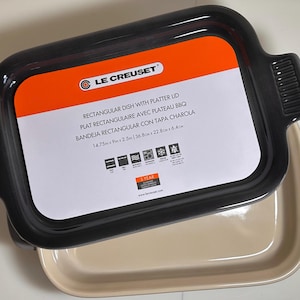 May include: A rectangular Le Creuset dish with a platter lid. The lid is black, and the dish is beige. The dimensions are 14.75 x 9 x 2.5 inches. The dish has handles on each side.