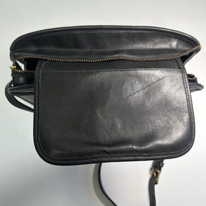 May include: A black leather shoulder bag featuring a top zipper closure and a front flap pocket. The bag has a structured rectangular shape with visible stitching along the edges and a thin adjustable shoulder strap with gold-tone metal hardware.