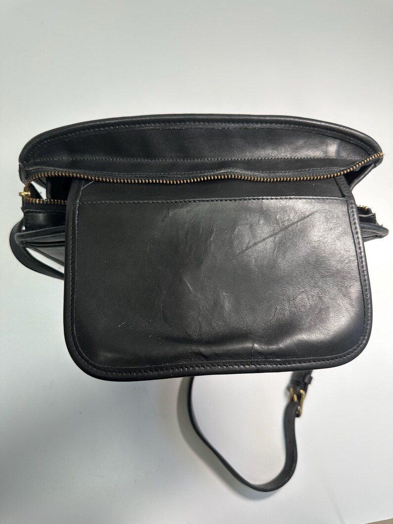 May include: A black leather shoulder bag featuring a top zipper closure and a front flap pocket. The bag has a structured rectangular shape with visible stitching along the edges and a thin adjustable shoulder strap with gold-tone metal hardware.
