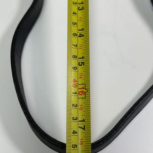 May include: A black leather strap positioned next to a yellow measuring tape. The tape shows the strap width is approximately 0.75 inches, while the visible length of the strap spans from the 12-inch mark to beyond the 18-inch mark.