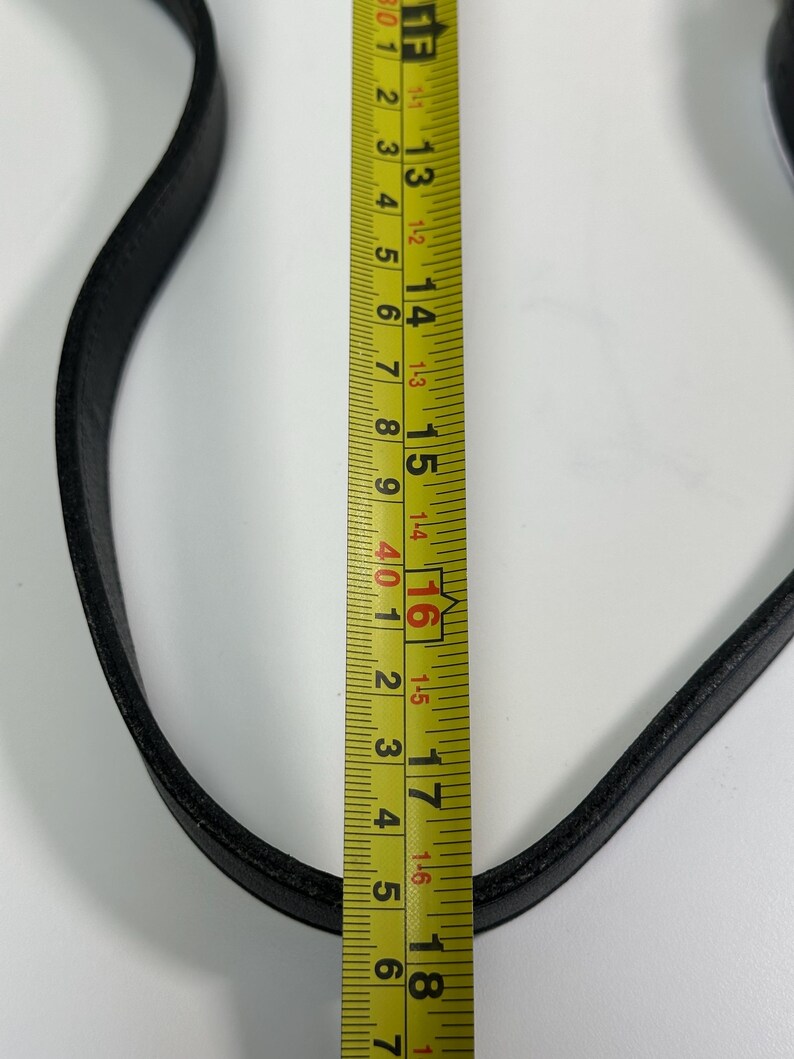 May include: A black leather strap positioned next to a yellow measuring tape. The tape shows the strap width is approximately 0.75 inches, while the visible length of the strap spans from the 12-inch mark to beyond the 18-inch mark.