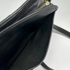 May include: A black leather shoulder bag featuring a smooth exterior finish and a top zipper closure with a gold-toned metal pull. The interior is lined with black fabric and includes a zippered pocket along the side panel with a matching metal pull.