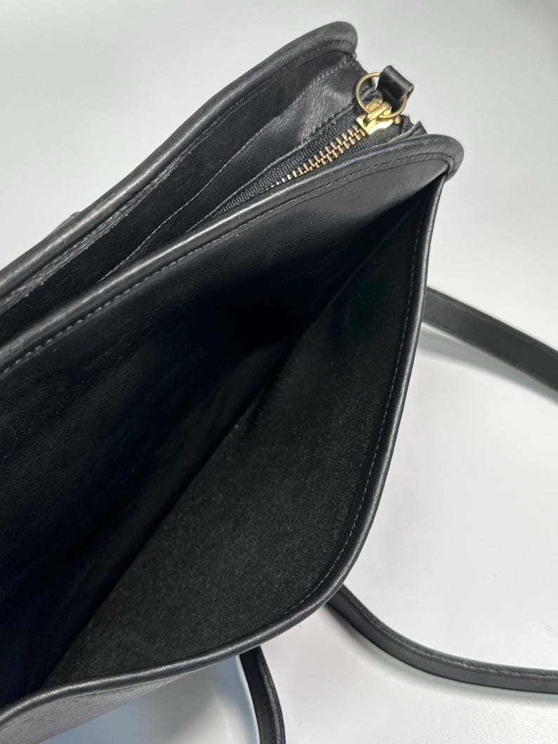 May include: A black leather shoulder bag featuring a smooth exterior finish and a top zipper closure with a gold-toned metal pull. The interior is lined with black fabric and includes a zippered pocket along the side panel with a matching metal pull.
