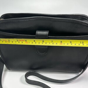 May include: A black shoulder bag with a smooth finish, featuring a front slip pocket and a central tab closure. A yellow measuring tape is positioned across the front, indicating a width of approximately 11.5 inches for the main body.