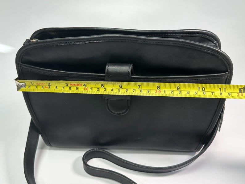 May include: A black shoulder bag with a smooth finish, featuring a front slip pocket and a central tab closure. A yellow measuring tape is positioned across the front, indicating a width of approximately 11.5 inches for the main body.