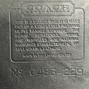 May include: A black leather surface featuring an embossed Coach Leatherware logo and a rectangular patch with the text: THIS IS A COACH BAG. IT IS MADE OUT OF A COMPLETELY NATURAL GLOVE TANNED COWHIDE. THE SCARS, SCRATCHES, VEINS, AND WRINKLES ARE NATURAL MARKINGS CHARACTERISTIC OF FULL-GRAIN LEATHERS. MADE IN THE UNITED STATES. NO 0486-229.