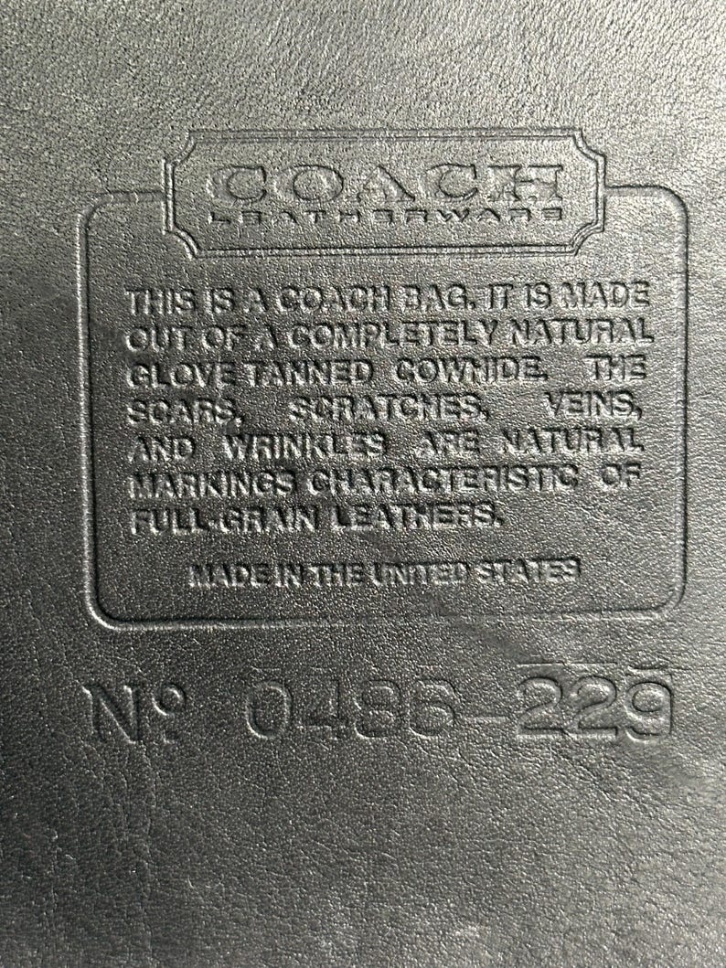 May include: A black leather surface featuring an embossed Coach Leatherware logo and a rectangular patch with the text: THIS IS A COACH BAG. IT IS MADE OUT OF A COMPLETELY NATURAL GLOVE TANNED COWHIDE. THE SCARS, SCRATCHES, VEINS, AND WRINKLES ARE NATURAL MARKINGS CHARACTERISTIC OF FULL-GRAIN LEATHERS. MADE IN THE UNITED STATES. NO 0486-229.