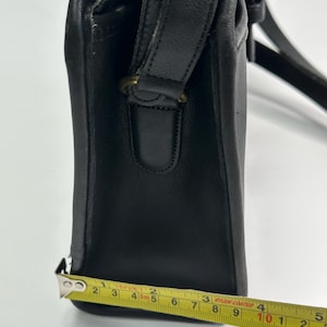 May include: A black leather shoulder bag with a side profile view. A yellow measuring tape is positioned along the base, showing a width of approximately 4.5 inches. The bag features a stitched leather strap attachment with a brass-toned metal ring.