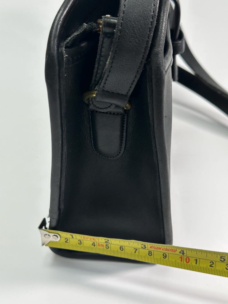 May include: A black leather shoulder bag with a side profile view. A yellow measuring tape is positioned along the base, showing a width of approximately 4.5 inches. The bag features a stitched leather strap attachment with a brass-toned metal ring.