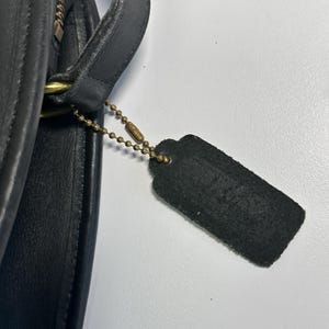 May include: A black leather handbag featuring a smooth texture and a curved edge. A rectangular black leather tag hangs from a brass-toned metal ball chain attached to the side of the bag near a brass-toned zipper and a leather strap attachment.