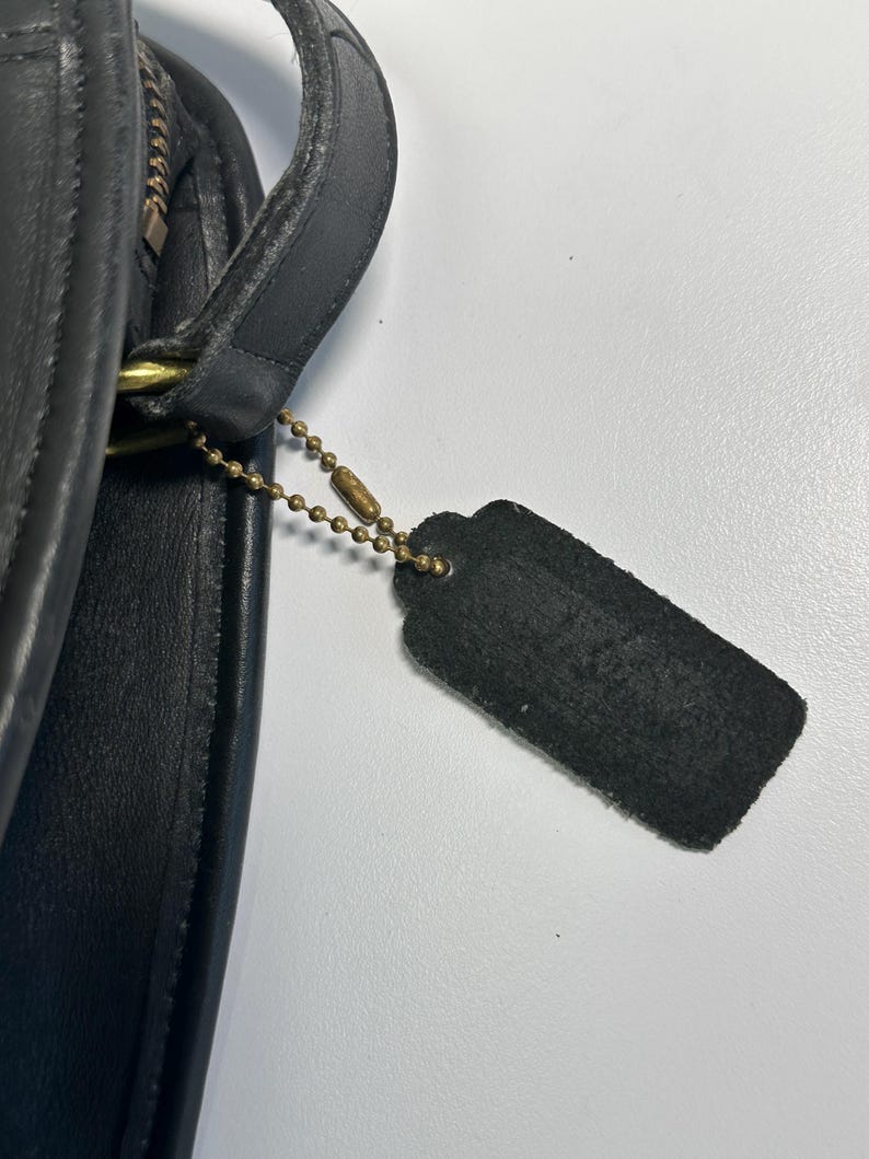 May include: A black leather handbag featuring a smooth texture and a curved edge. A rectangular black leather tag hangs from a brass-toned metal ball chain attached to the side of the bag near a brass-toned zipper and a leather strap attachment.