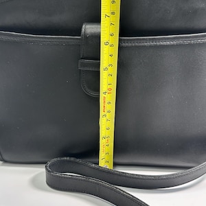 May include: A black leather shoulder bag with a front flap closure and a long strap. A yellow measuring tape is placed vertically over the center, showing a height of approximately 8 inches from the bottom to the top edge of the bag.