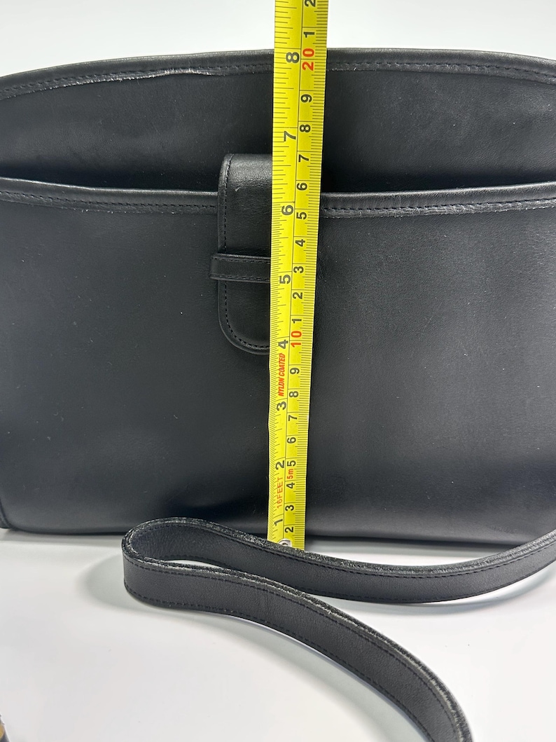 May include: A black leather shoulder bag with a front flap closure and a long strap. A yellow measuring tape is placed vertically over the center, showing a height of approximately 8 inches from the bottom to the top edge of the bag.