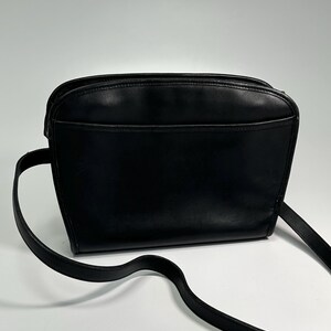May include: A black leather crossbody bag with a smooth finish, featuring a rounded top silhouette, a zippered main compartment, and a thin, adjustable shoulder strap. The front of the bag includes a horizontal slip pocket across the width.