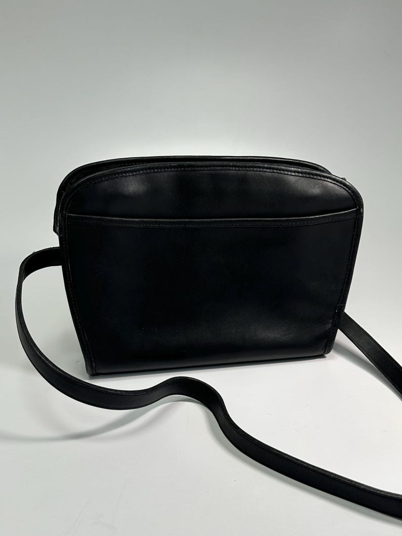 May include: A black leather crossbody bag with a smooth finish, featuring a rounded top silhouette, a zippered main compartment, and a thin, adjustable shoulder strap. The front of the bag includes a horizontal slip pocket across the width.