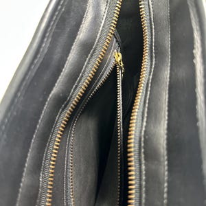May include: A black leather handbag interior featuring a main compartment with a gold-toned metal zipper closure. The interior lining is black, and there is an additional zippered pocket on the side with a small gold-toned metal zipper pull.