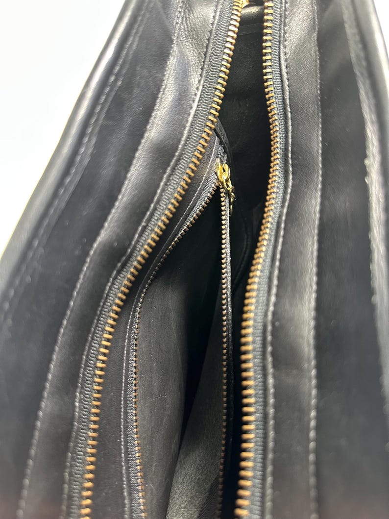 May include: A black leather handbag interior featuring a main compartment with a gold-toned metal zipper closure. The interior lining is black, and there is an additional zippered pocket on the side with a small gold-toned metal zipper pull.