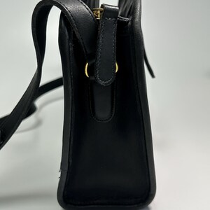 May include: A black leather shoulder bag with a structured rectangular profile, featuring a top zipper closure with a gold-toned metal pull, a matching black leather strap, and gold-toned hardware rings connecting the strap to the bag body.
