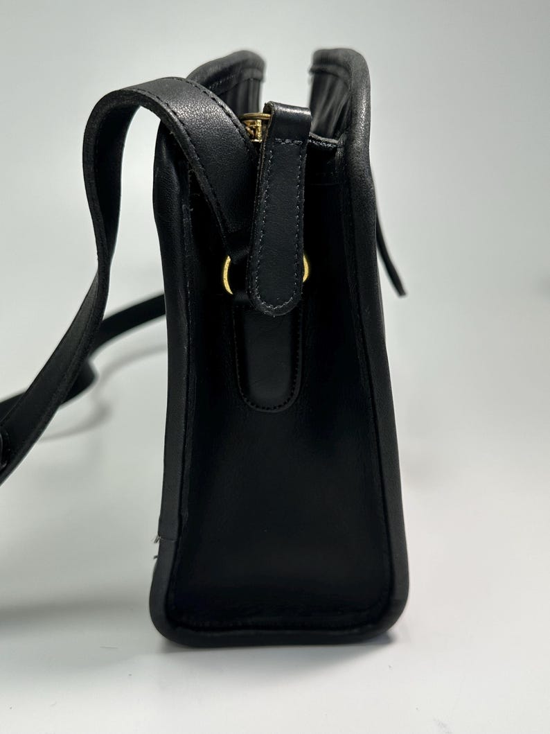 May include: A black leather shoulder bag with a structured rectangular profile, featuring a top zipper closure with a gold-toned metal pull, a matching black leather strap, and gold-toned hardware rings connecting the strap to the bag body.