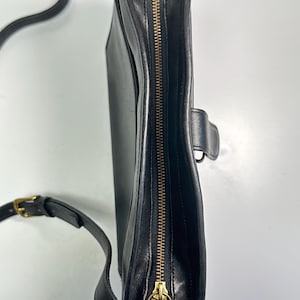 May include: A black leather crossbody bag viewed from the side, featuring a brass-toned metal zipper closure along the top edge, a matching brass-toned buckle on the adjustable shoulder strap, and a small side tab with a metal ring attachment.