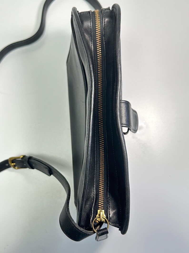 May include: A black leather crossbody bag viewed from the side, featuring a brass-toned metal zipper closure along the top edge, a matching brass-toned buckle on the adjustable shoulder strap, and a small side tab with a metal ring attachment.