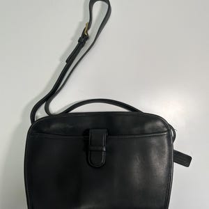 May include: A black leather shoulder bag featuring a top handle, an adjustable shoulder strap with a brass buckle, and a front exterior pocket secured by a vertical leather tab closure. A small leather hang tag is attached to the side of the bag.