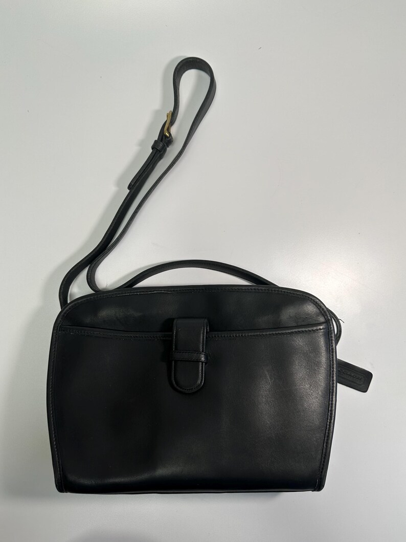 May include: A black leather shoulder bag featuring a top handle, an adjustable shoulder strap with a brass buckle, and a front exterior pocket secured by a vertical leather tab closure. A small leather hang tag is attached to the side of the bag.