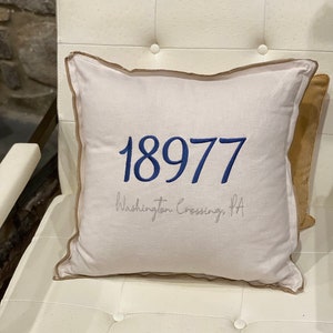 Könnte beinhalten: Ein weißes Kissen mit braunem Besatz und blauer Stickerei. Das Kissen zeigt die Nummer "18977" und den Text "Washington Crossing, PA".