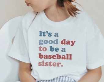 May include: A white t-shirt with the text "it's a good day to be a baseball sister" in blue and red. The shirt is worn by a child, paired with striped pants and red sneakers. The child is wearing white sunglasses.