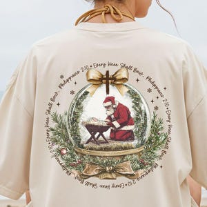 May include: Cream-colored oversized t-shirt featuring a Christmas-themed graphic on the back. The graphic depicts Santa Claus kneeling before a manger inside a snow globe, surrounded by text and festive elements. The shirt is worn by a person sitting on a beach.