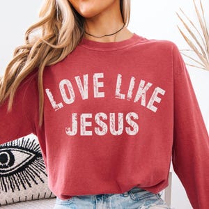 May include: A long-sleeve crewneck sweatshirt in a faded crimson red color. The front features the white distressed-style text "LOVE LIKE JESUS" printed in a bold, arched font across the chest. The garment has a relaxed fit and long sleeves.