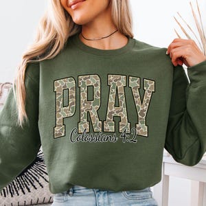 May include: Military green sweatshirt with the word "PRAY" in a camouflage pattern across the chest. Below the word "PRAY" is the text "Colossians 4:2". The sweatshirt is a crew neck style.